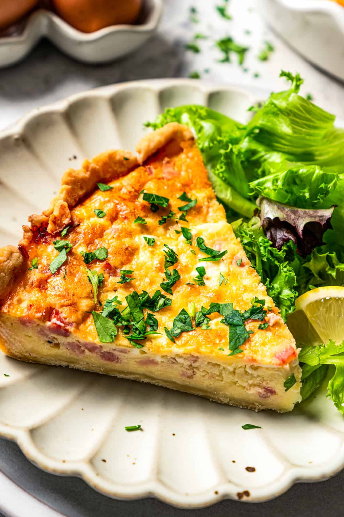 A slice of ham and cheese quiche next to a side of green salad on a scalloped white plate.