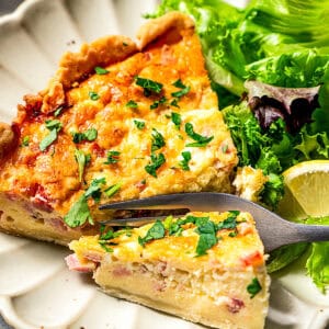 A fork cutting into a slice of quiche next to a side of green salad on a scalloped white plate.