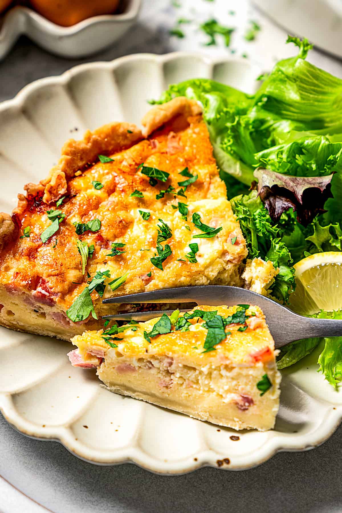 A fork cutting into a slice of quiche next to a side of green salad on a scalloped white plate.