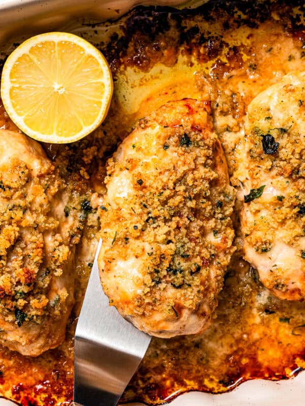 Overhead view of a spatula lifting up a parmesan crusted chicken breast next to more chicken breasts and lemon slices in a baking dish.