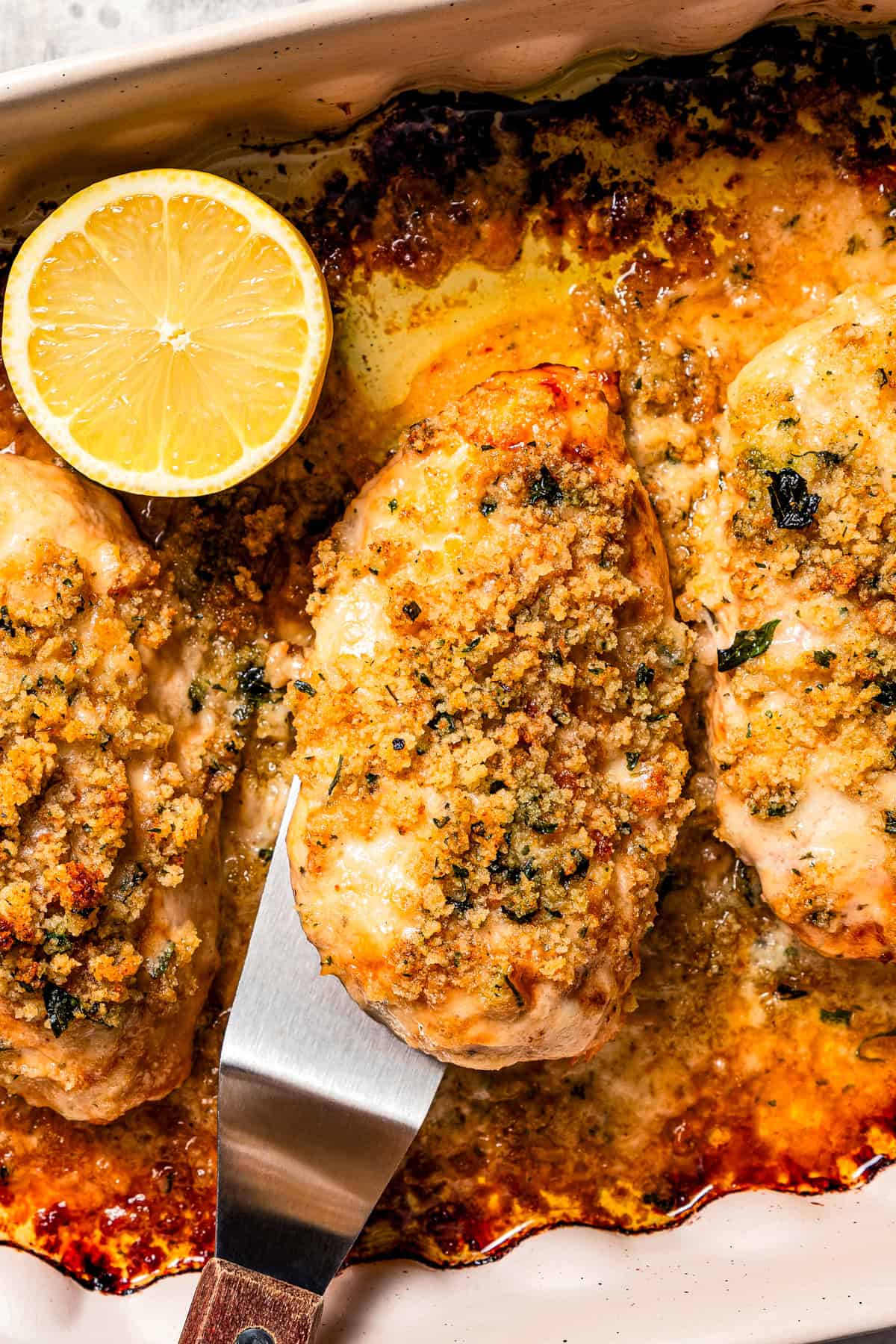 Overhead view of a spatula lifting up a parmesan crusted chicken breast next to more chicken breasts and lemon slices in a baking dish.