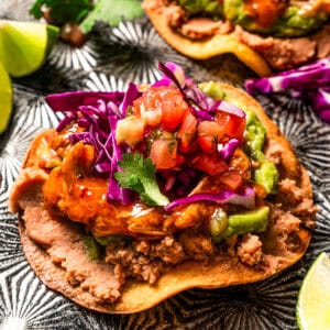 Close up of tostadas de tinga piled with toppings next to lime wedges on a baking sheet.