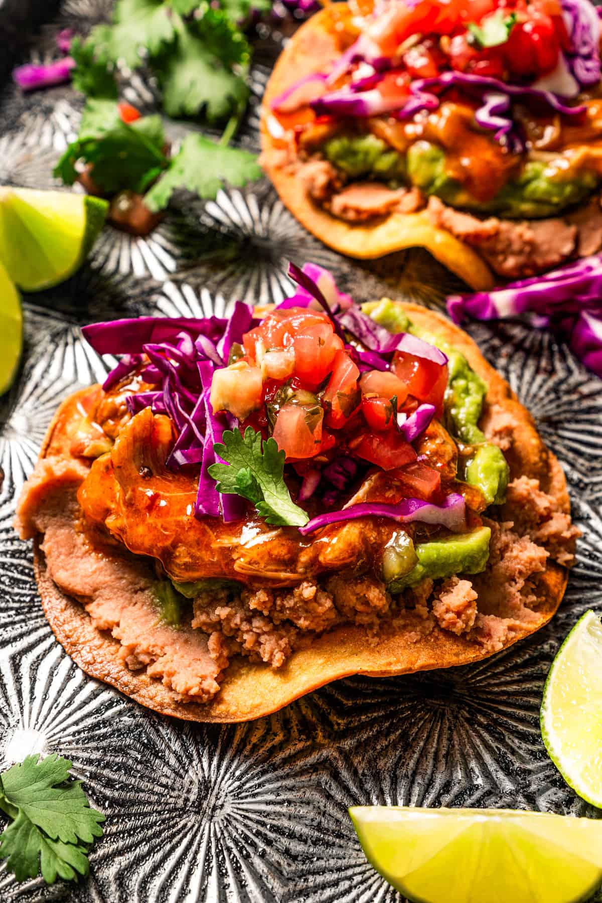 Close up of tostadas de tinga piled with toppings next to lime wedges on a baking sheet.