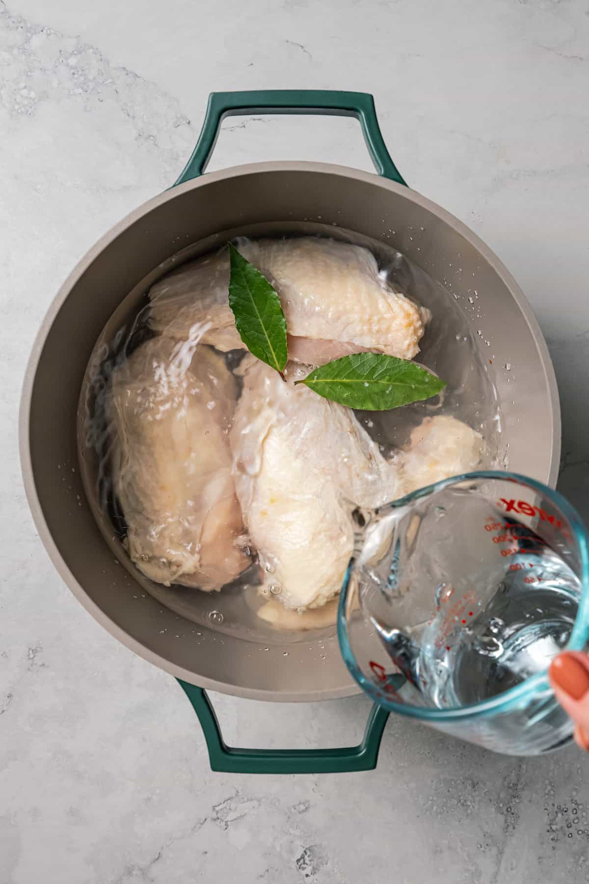 Water being poured into a pot with chicken breasts and bay leaves.