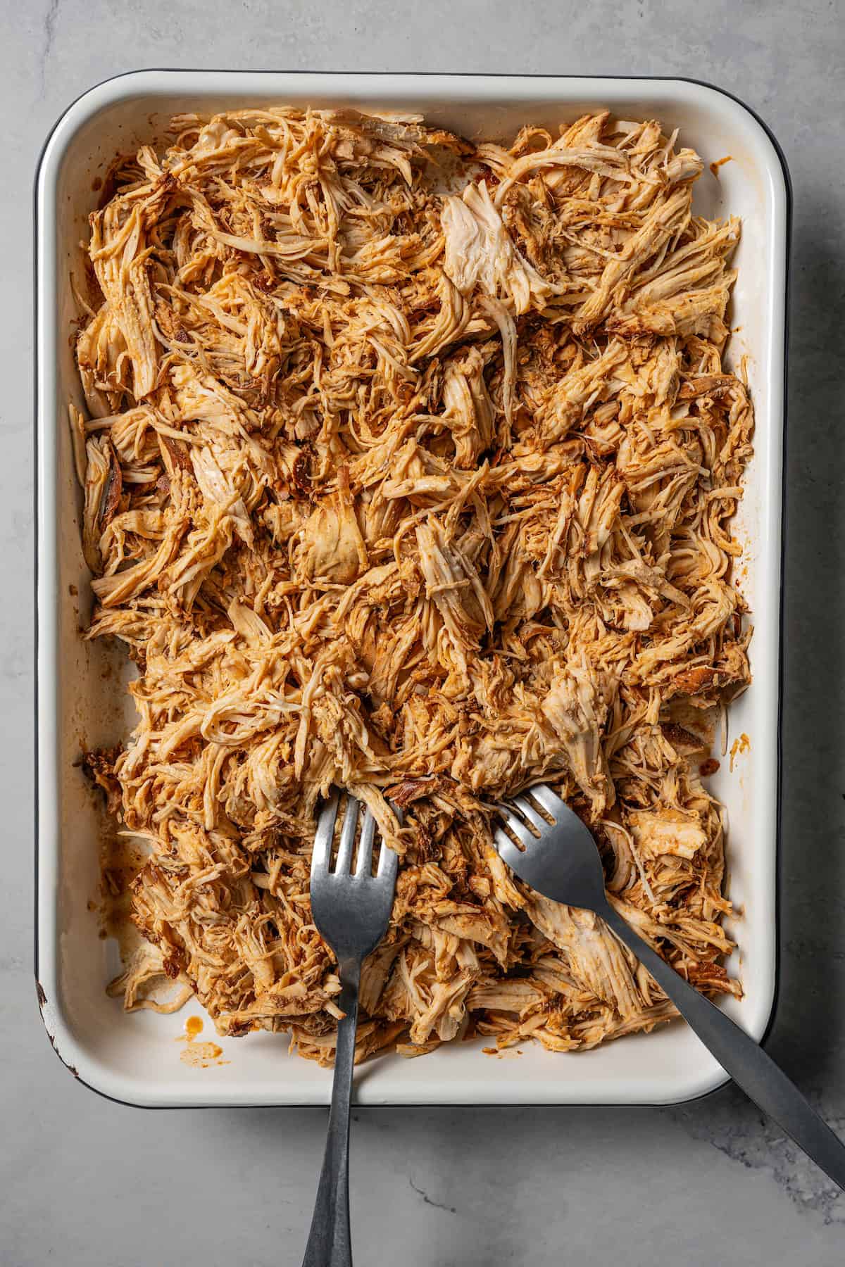 Two forks resting in a baking tray of shredded chicken.