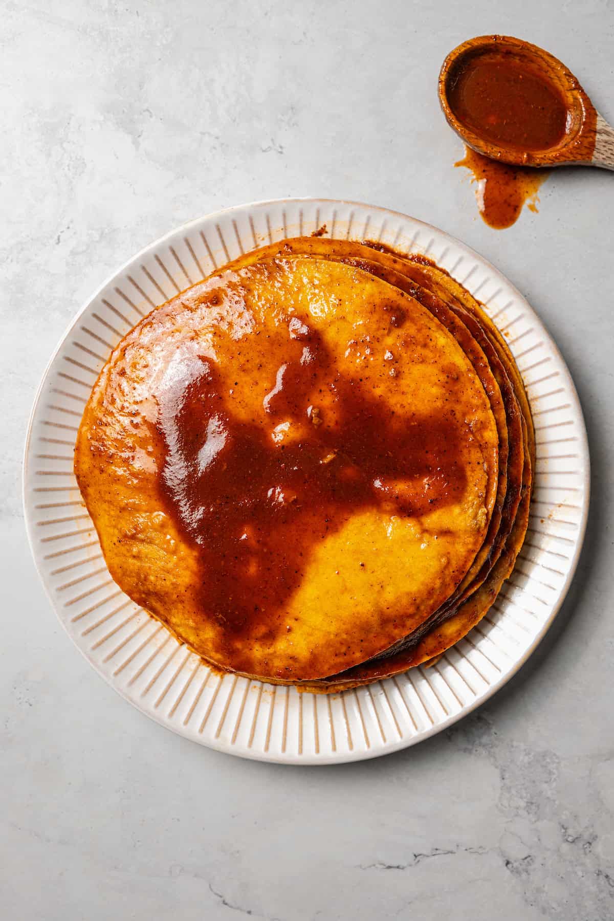 Overhead view of a stack of tortillas dipped in consomme on a plate next to a spoon with sauce dripping from the side.