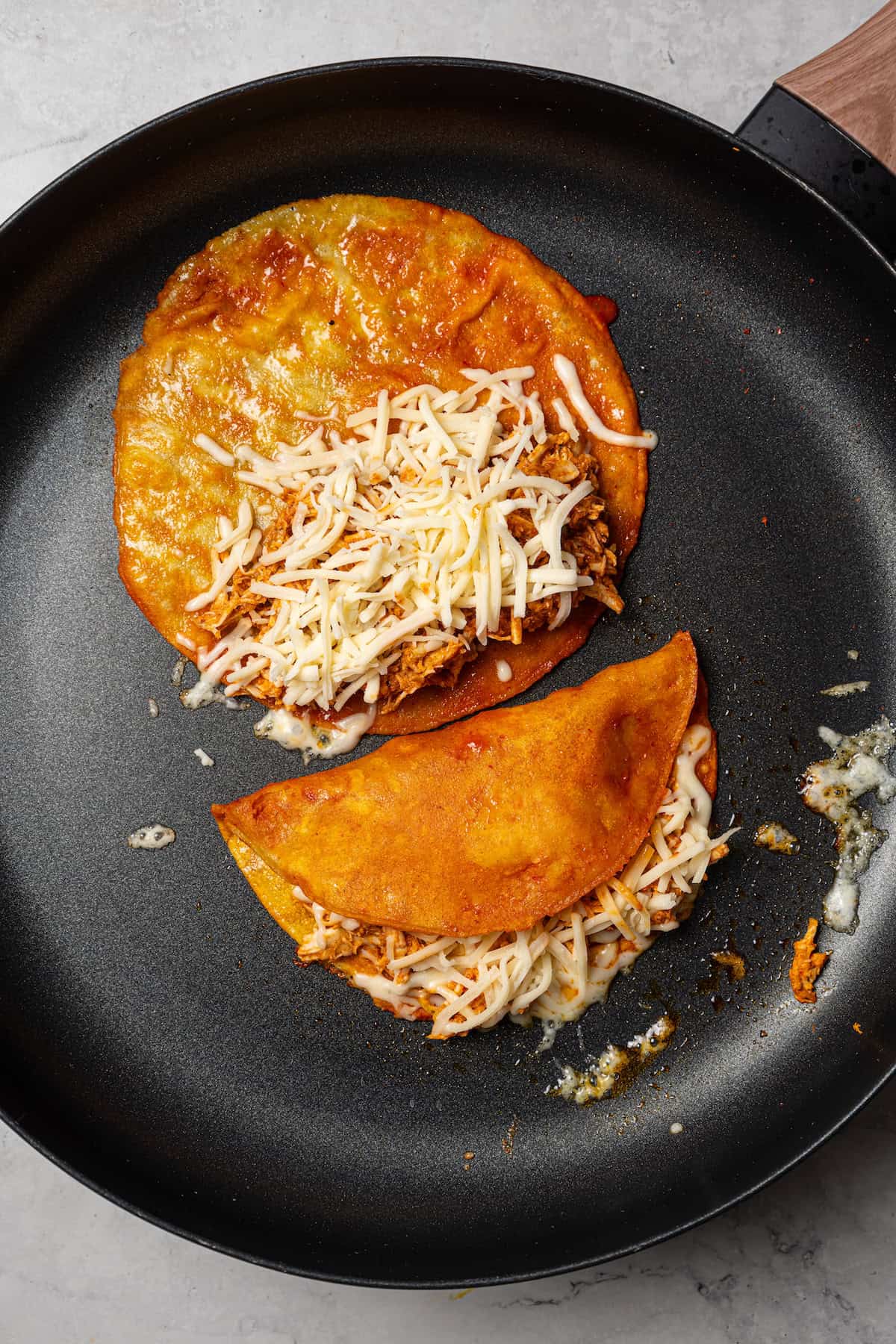 Two chicken birria tacos cooking in a skillet, one folded in half and the second topped with shredded cheese.