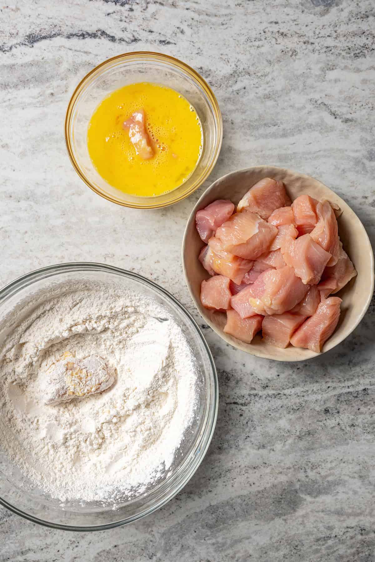 A bowl of uncooked chicken pieces next to a second bowl of cornstarch and a third bowl with beaten egg.