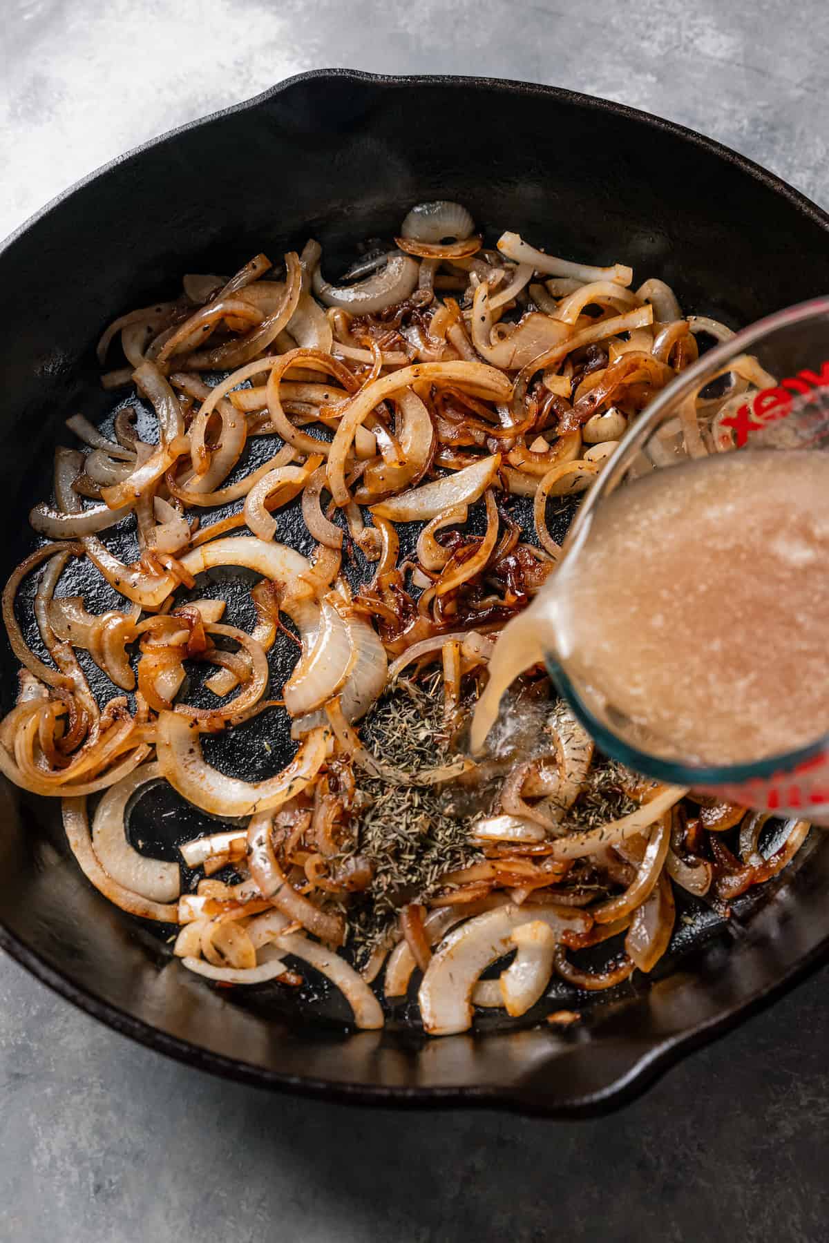 Beef broth and seasonings being poured over caramelized onions in a skillet.