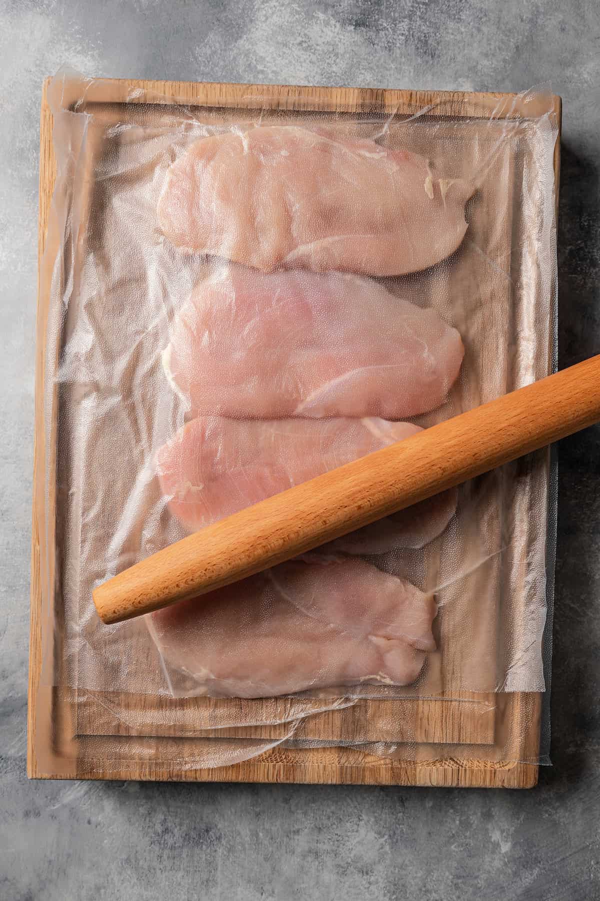 Chicken breasts pounded flat between two sheets of plastic wrap on a wooden cutting board, with a rolling pin resting on top.