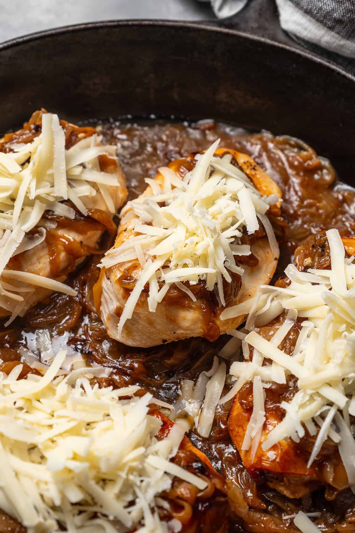 Chicken breasts topped with caramelized onions and shredded cheese in a skillet of French onion soup.