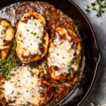 A skillet with four chicken breasts topped with caramelized onions and melted cheese.