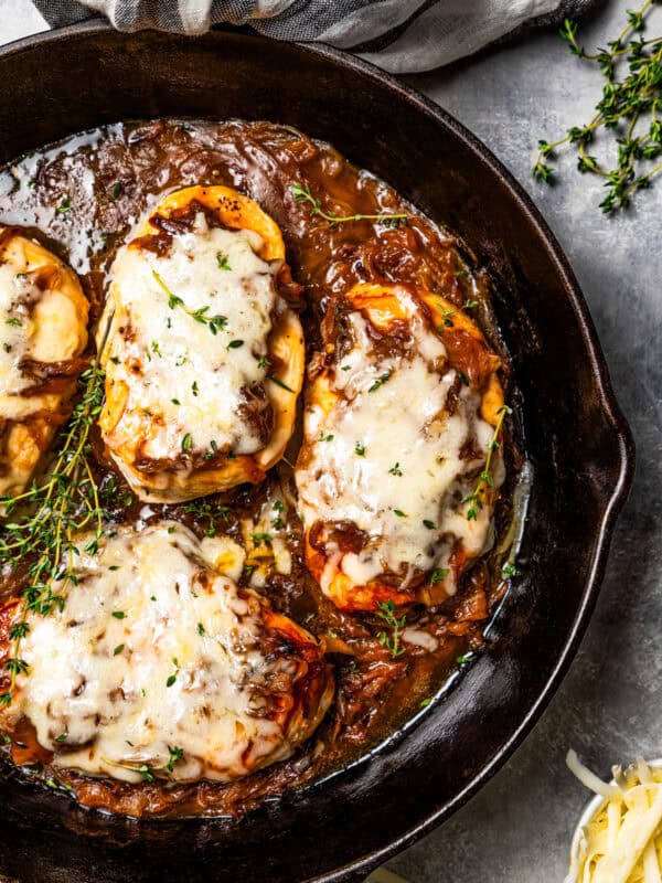 A skillet with four chicken breasts topped with caramelized onions and melted cheese.