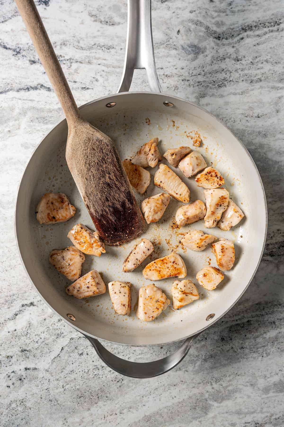 A wooden spoon stirring seared cubed chicken in a skillet.