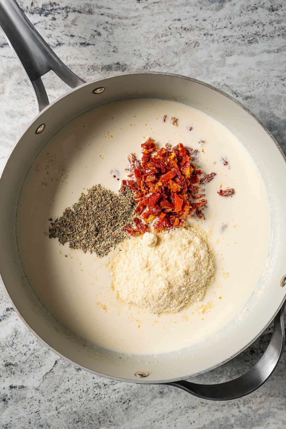 Sun-dried tomatoes, seasoning, and parmesan added to cream sauce in a skillet.