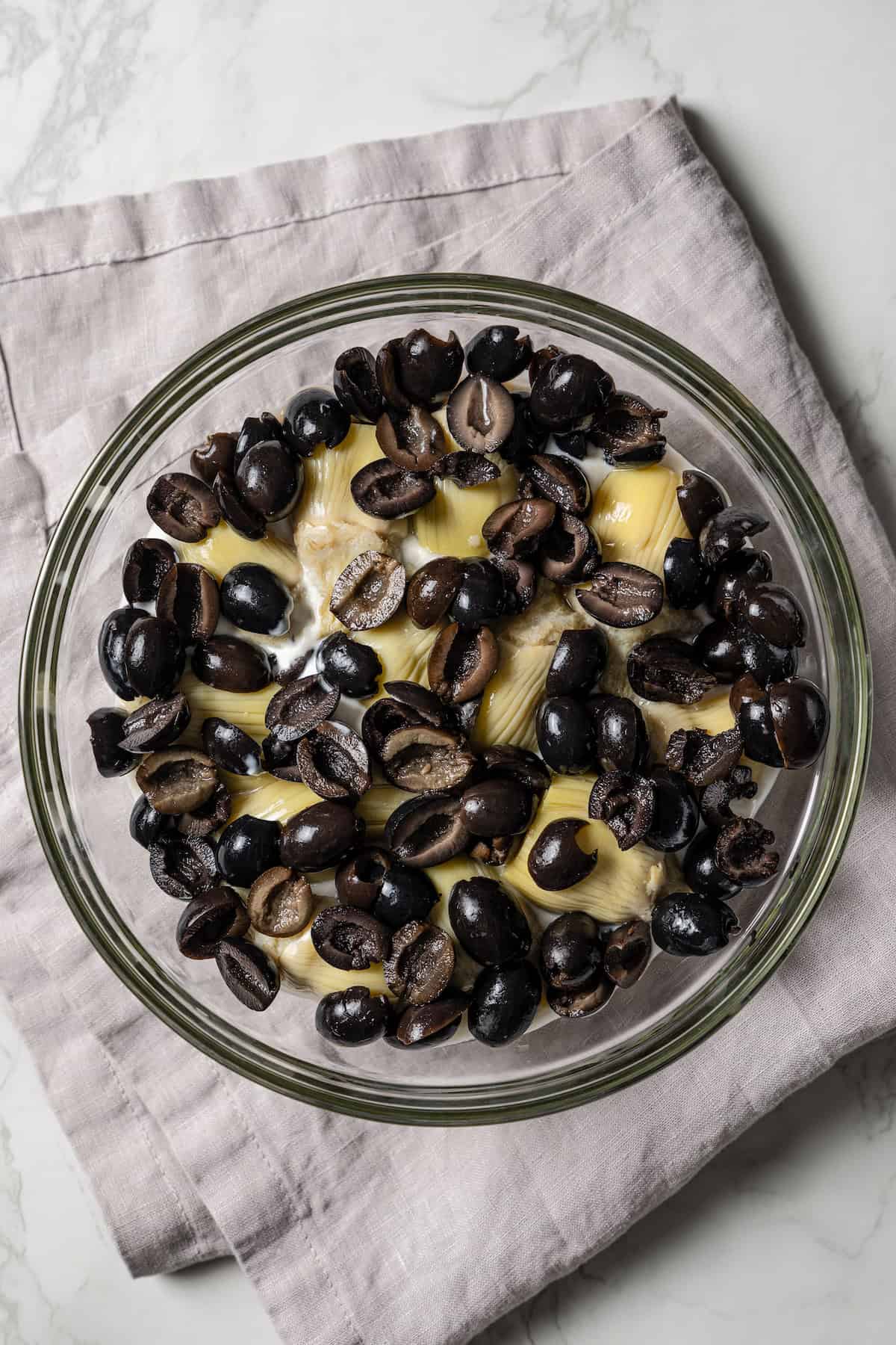 Olives layered over artichokes and dip ingredients in a glass bowl.