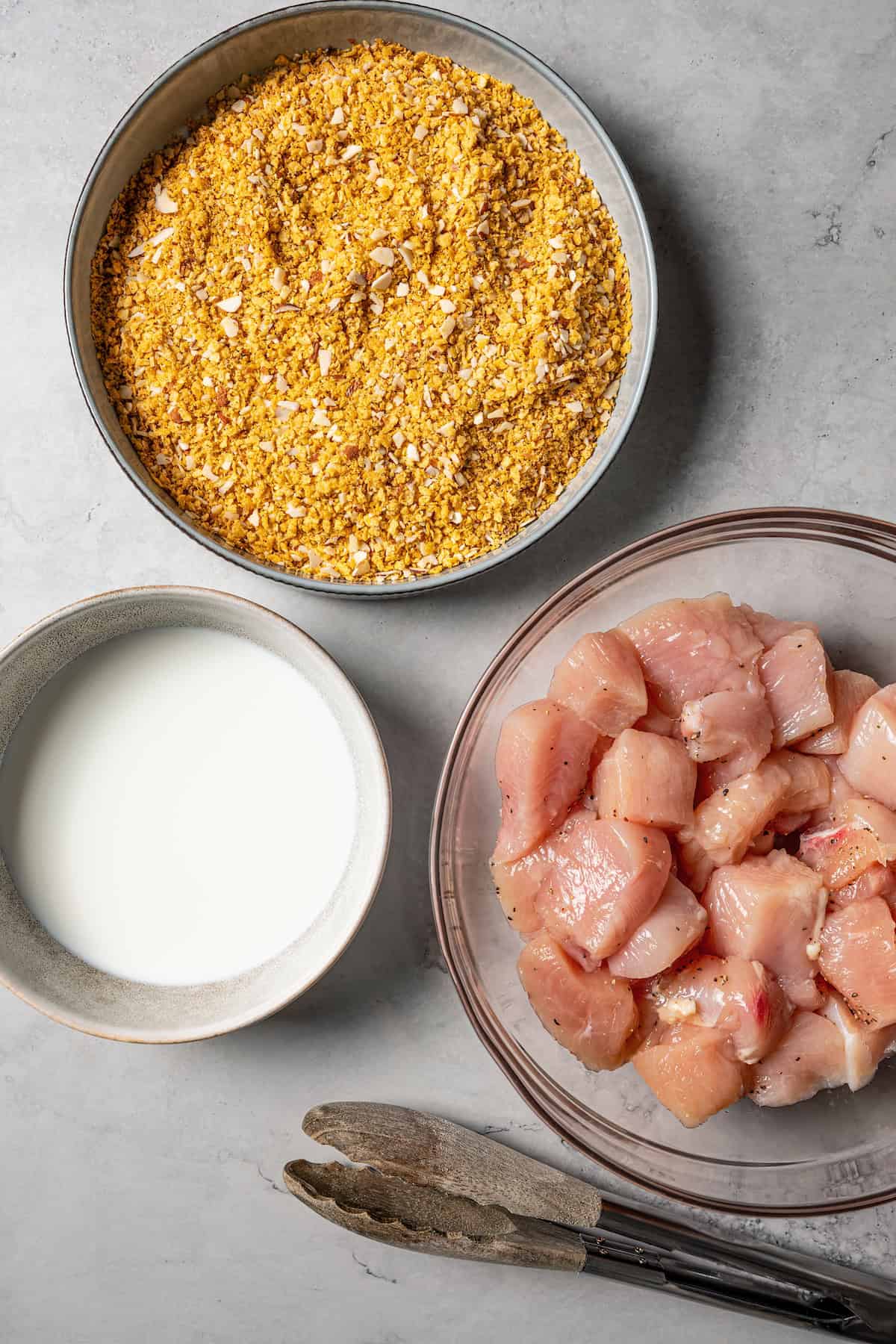 A breading station with a bowl of chicken, another bowl of crumbs, and a third bowl of milk.