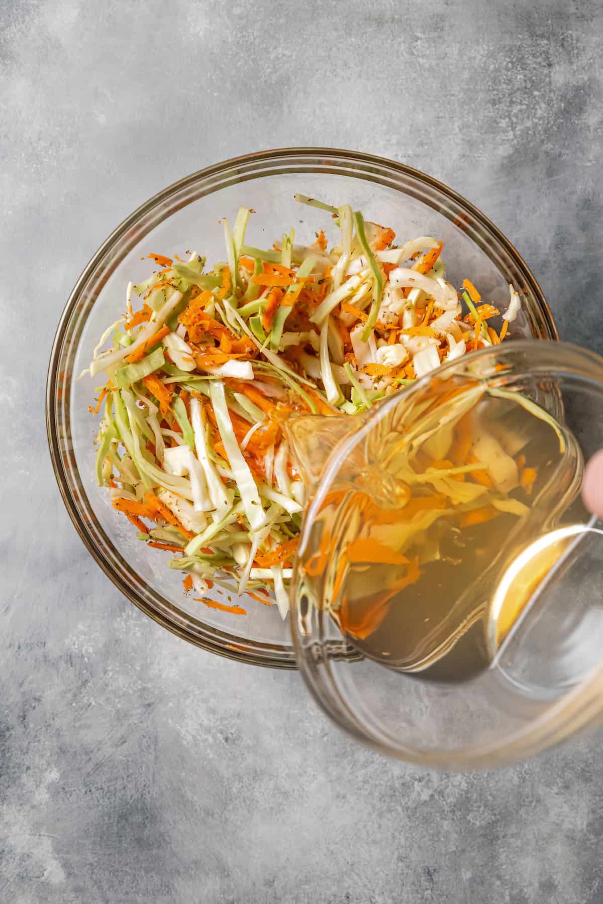 Vinegar dressing being poured over cabbage slaw in a glass bowl.