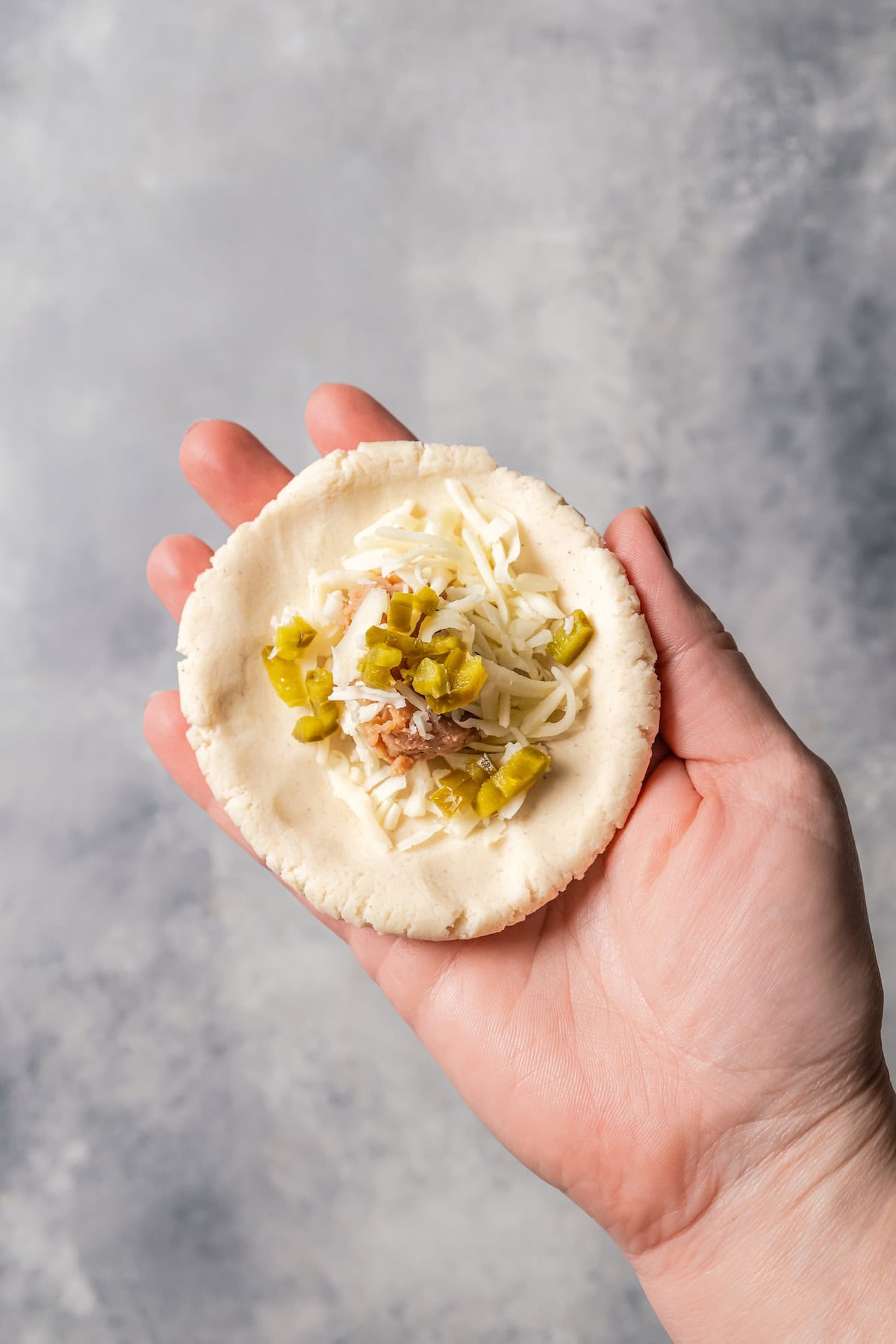 A hand holding flattened pupusa dough filled with shredded cheese and diced jalapeños.