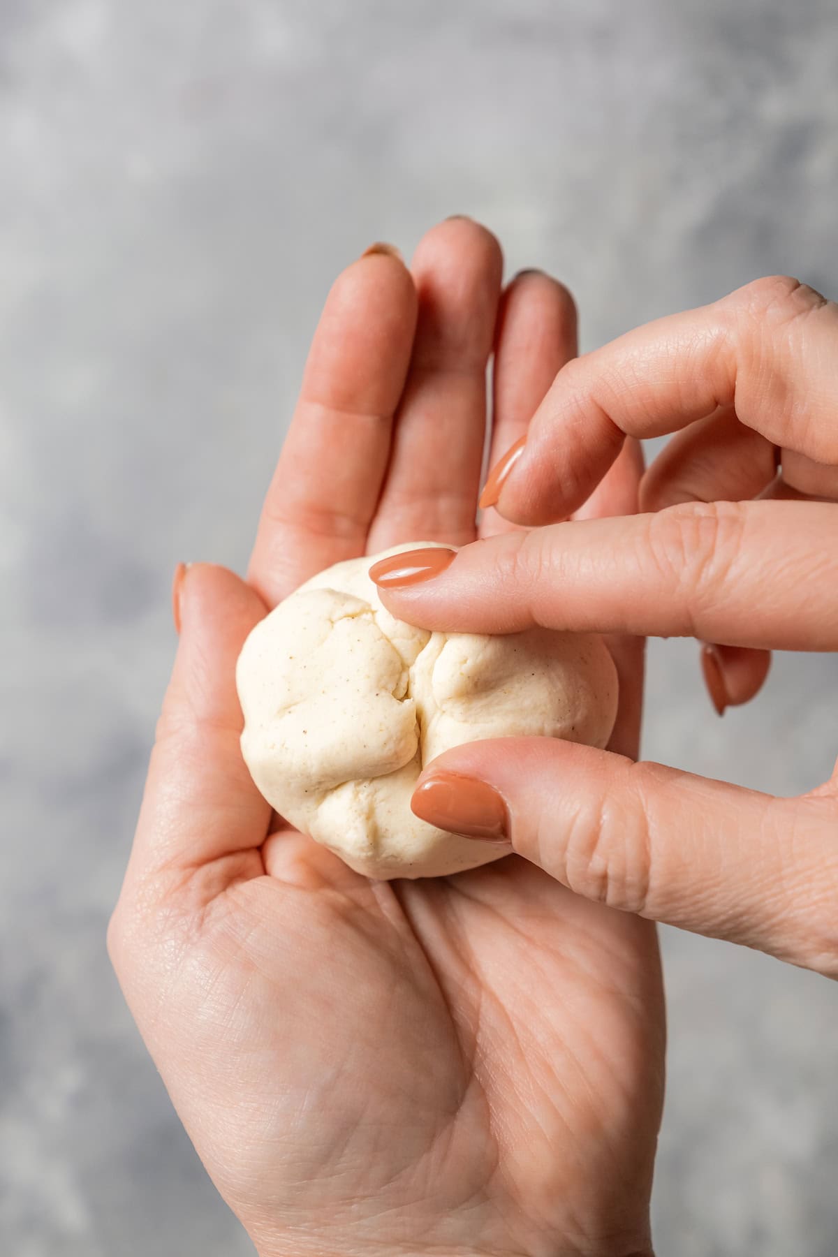 One hand holding a filled and folded pupusa dough ball while the other hand pinches the dough closed.