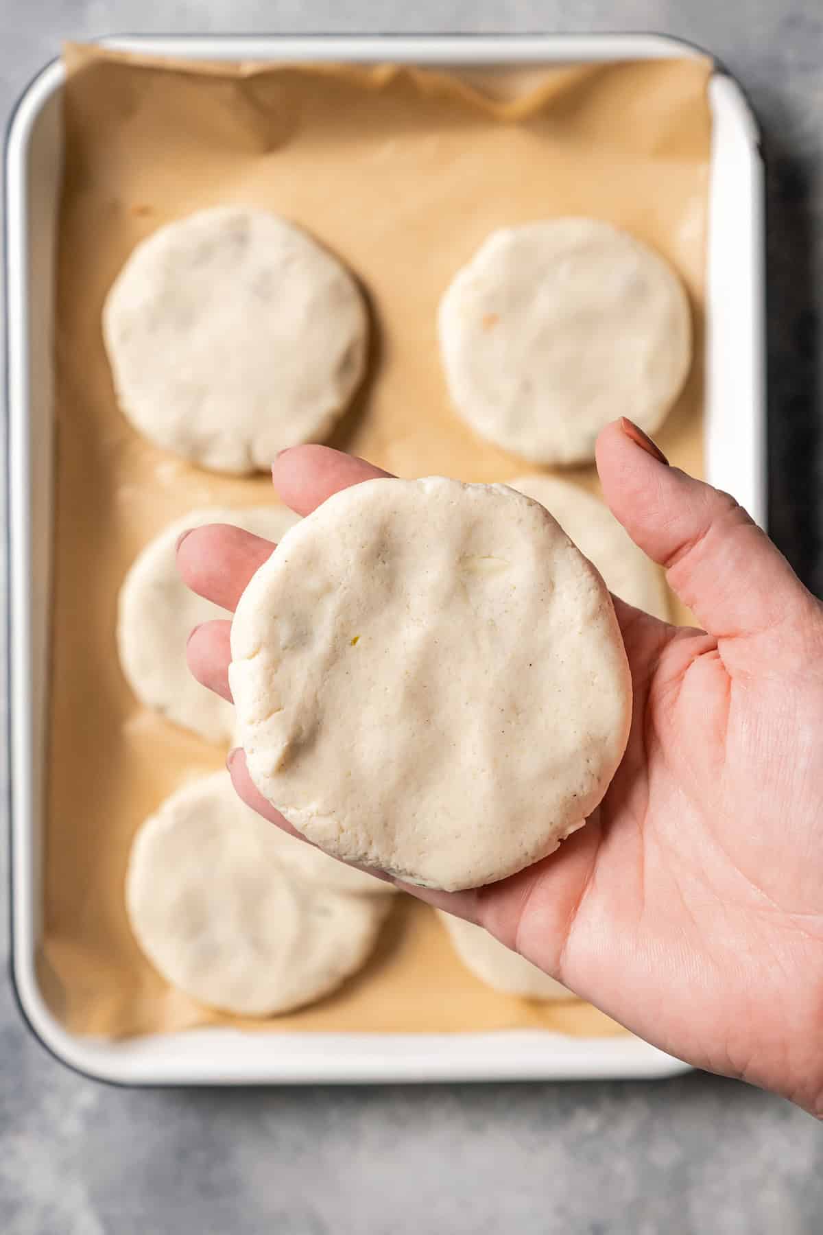 Close up of a hand holding up a filled and flattened pupusa, with more pupusas on a parchment-lined tray below.