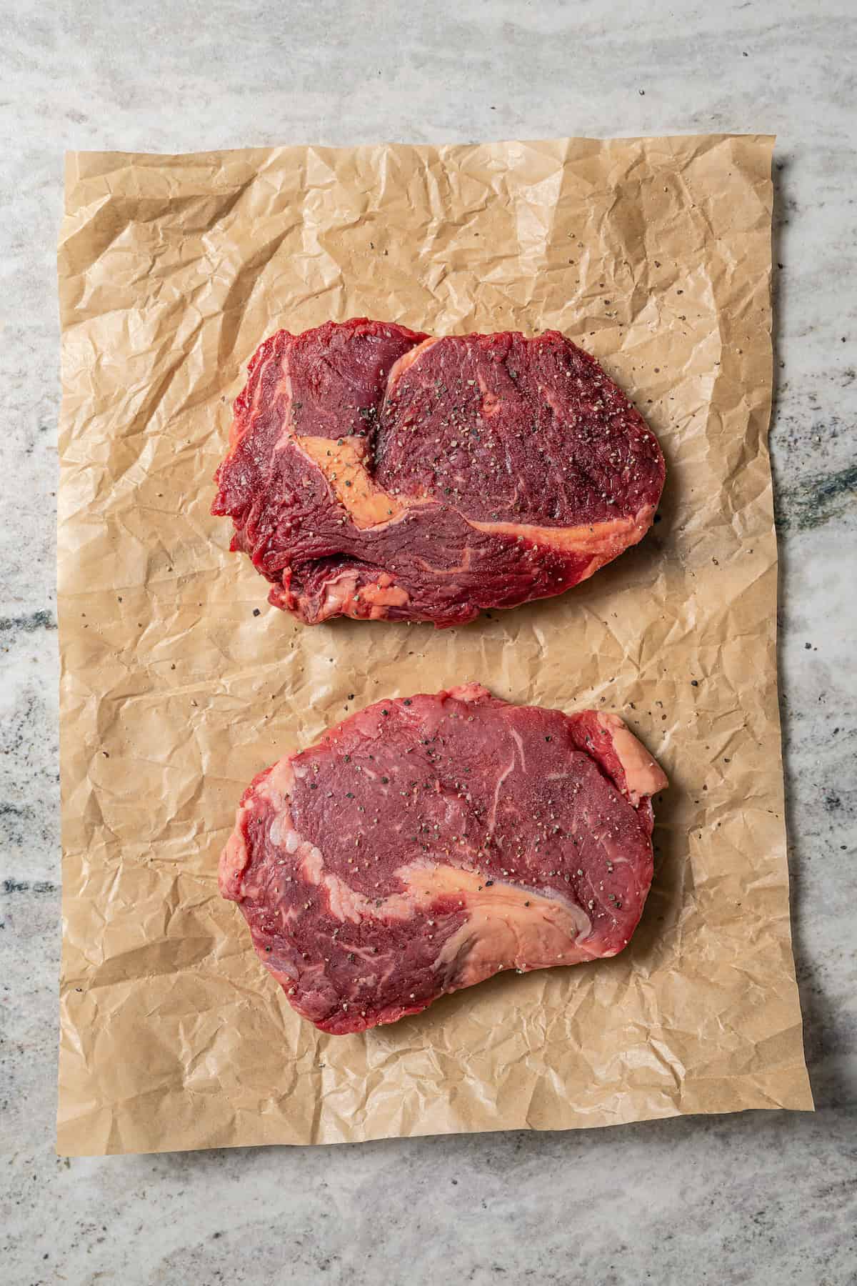 Two uncooked seasoned steaks on a piece of parchment paper.