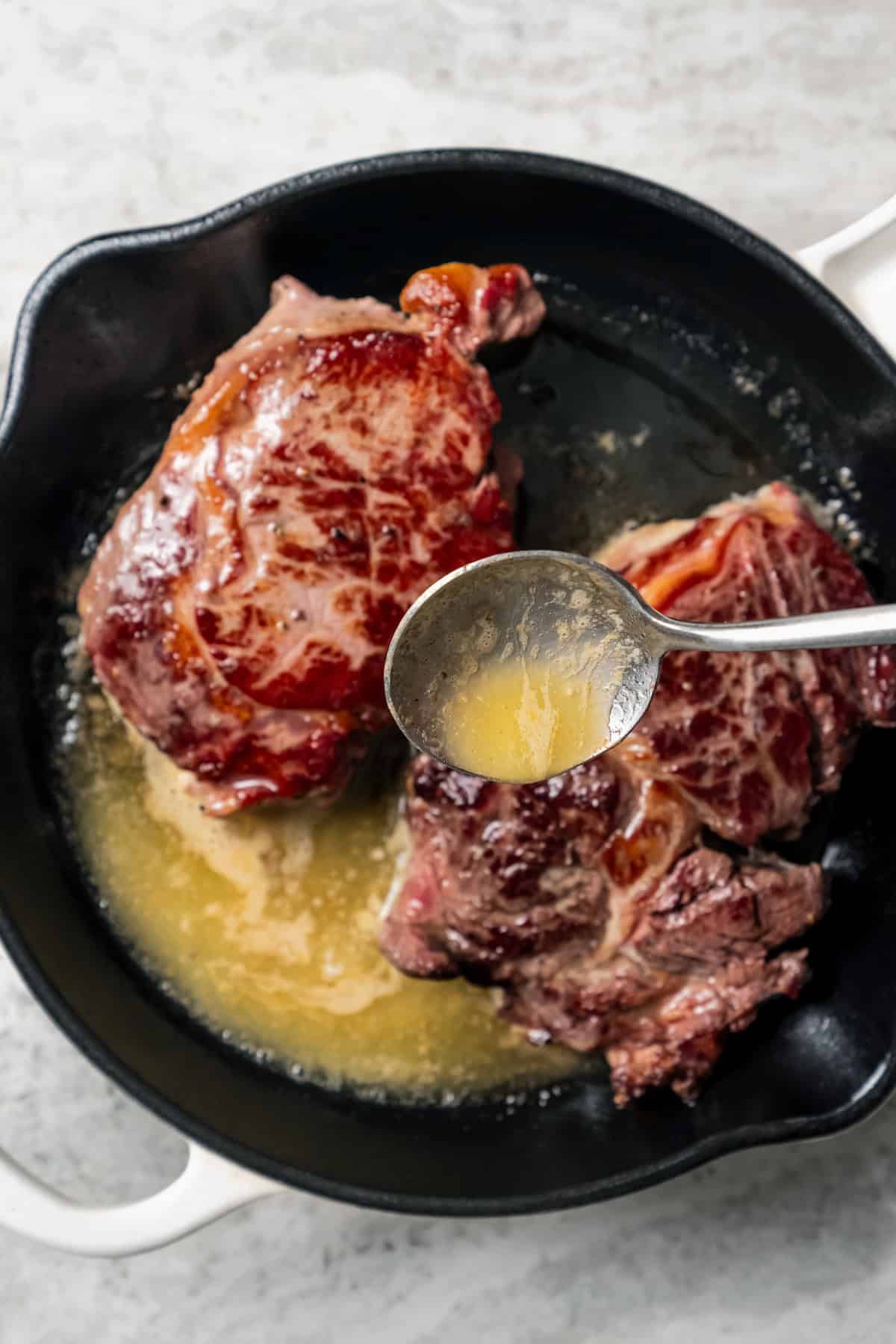 A spoon is used to baste two seared steaks with butter in a cast iron skillet.