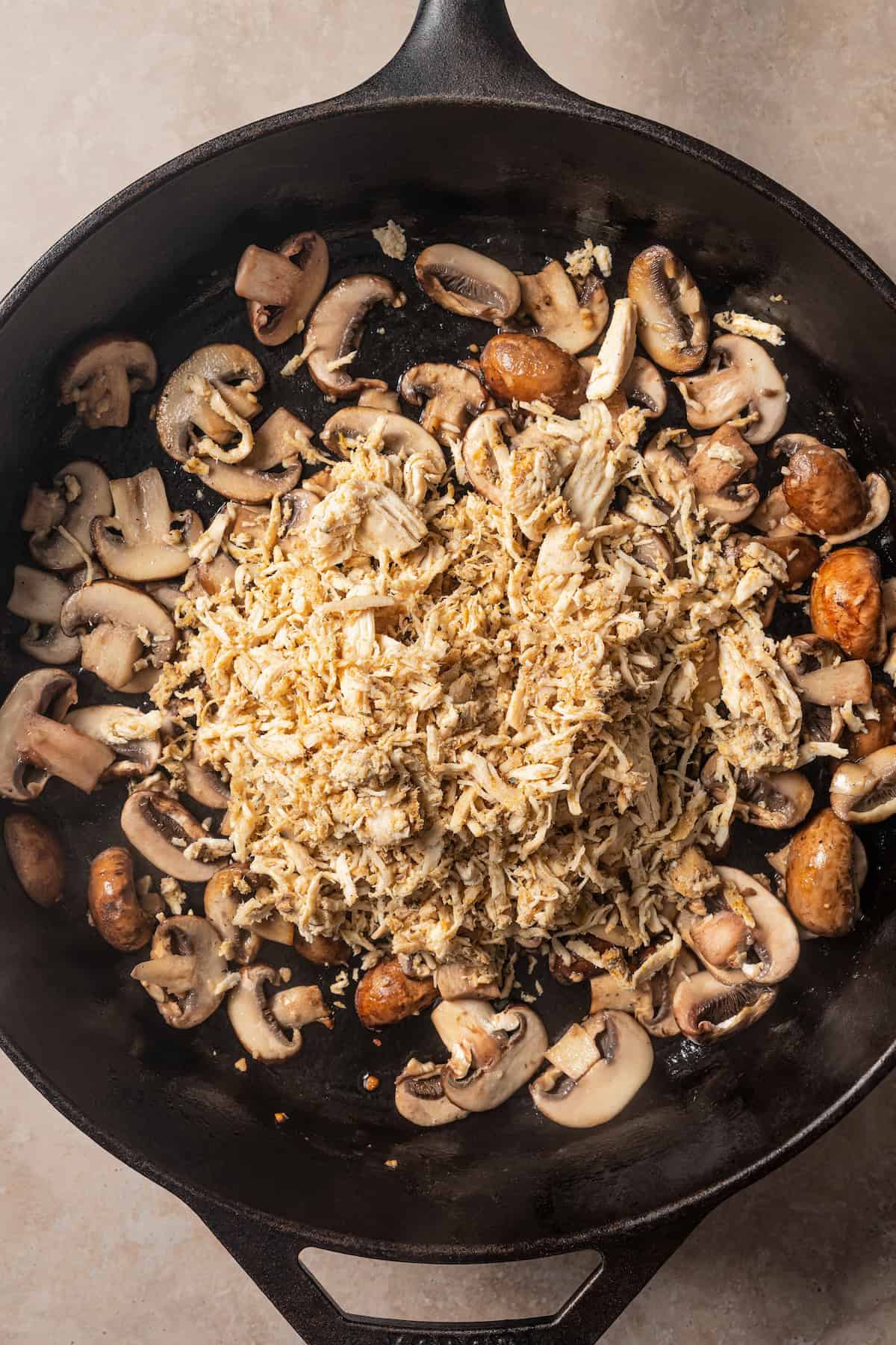 Shredded chicken added to a skillet with sautéed garlic mushrooms.