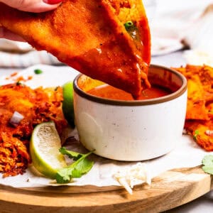 A hand dipping a chicken birria taco into a bowl of consomme on a wooden platter.