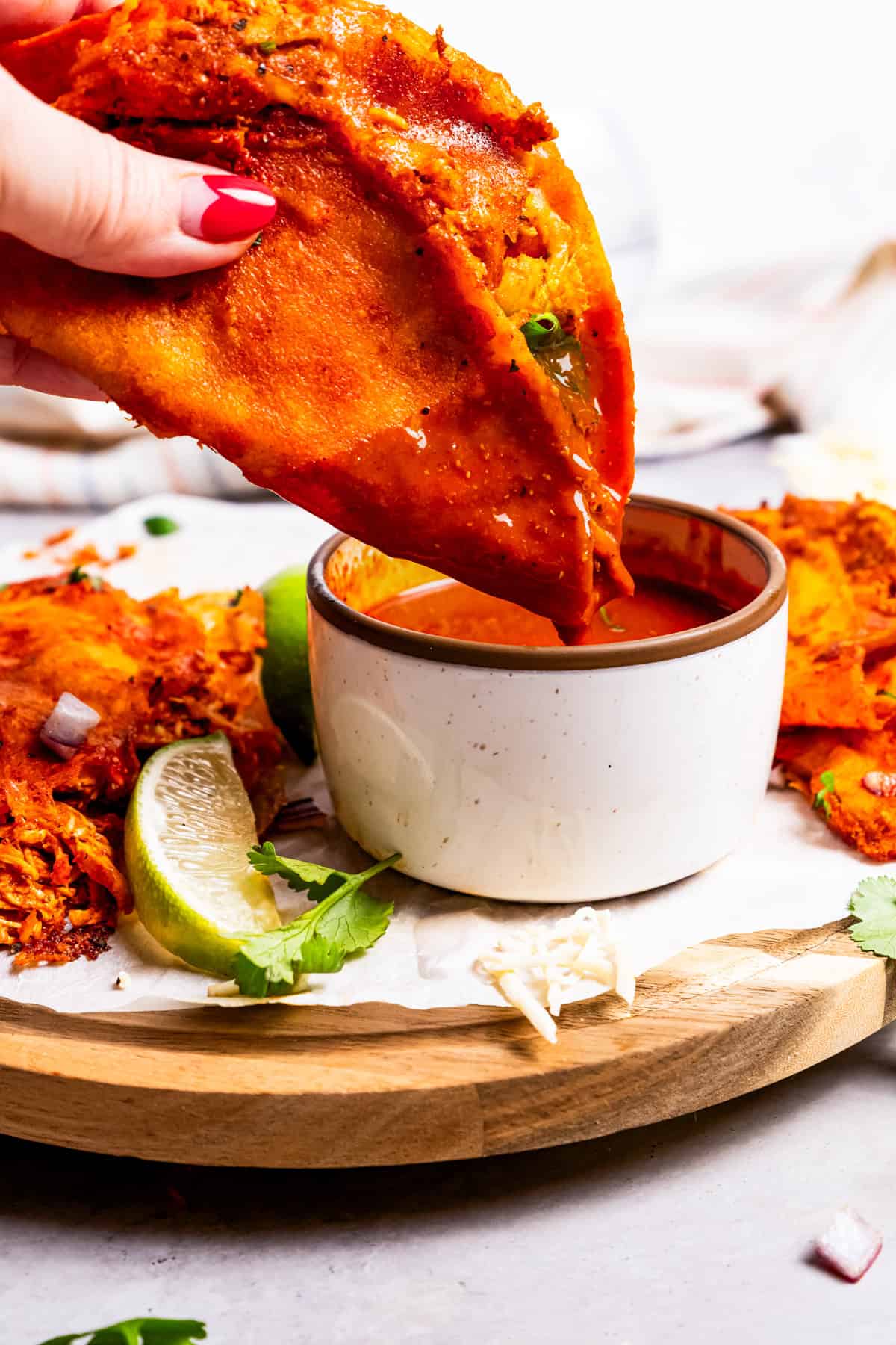 A hand dipping a chicken birria taco into a bowl of consomme on a wooden platter.