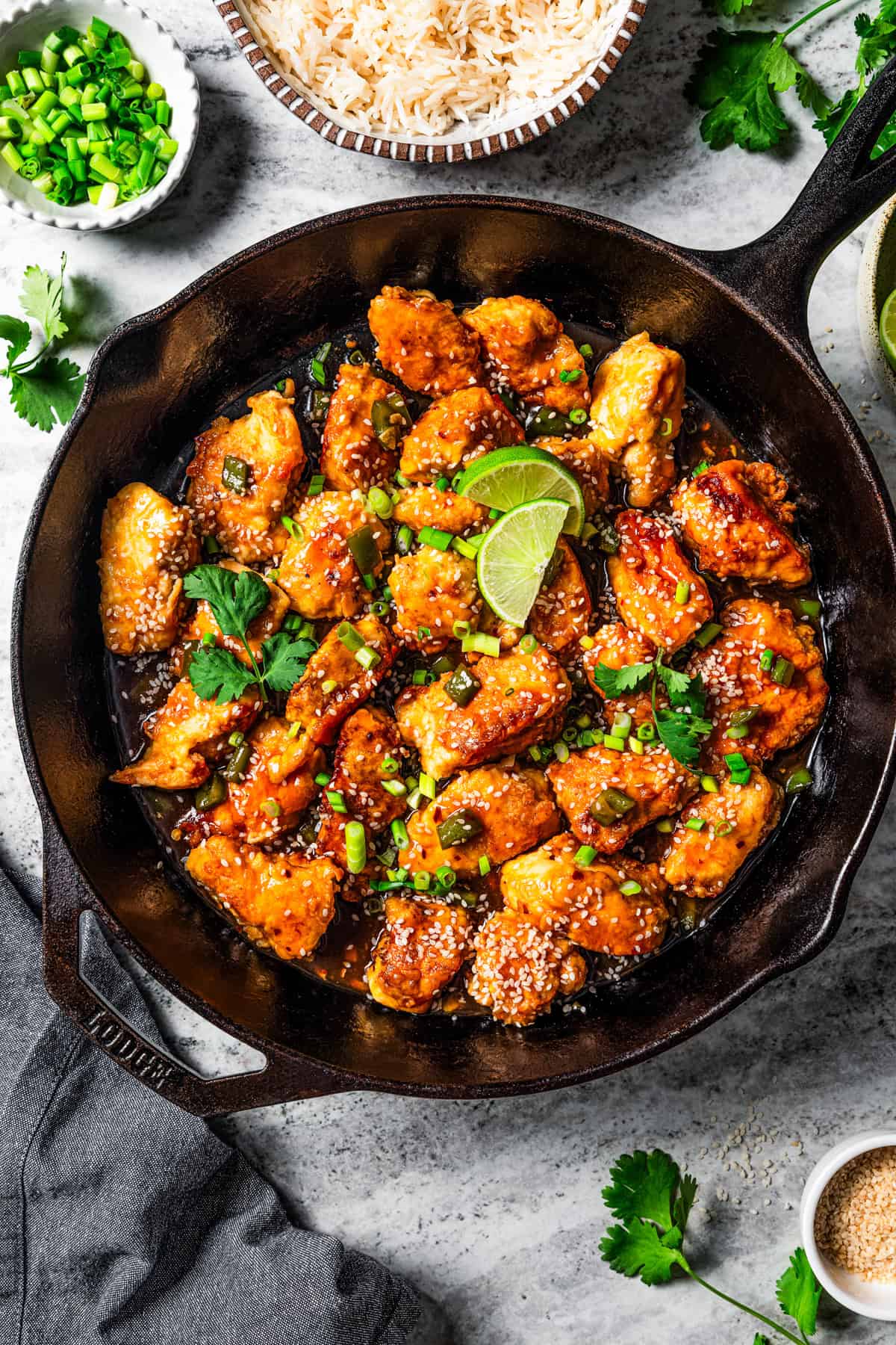 Overhead image of empress chicken in a skillet garnished with lime wedges, sesame seeds, and green onions.