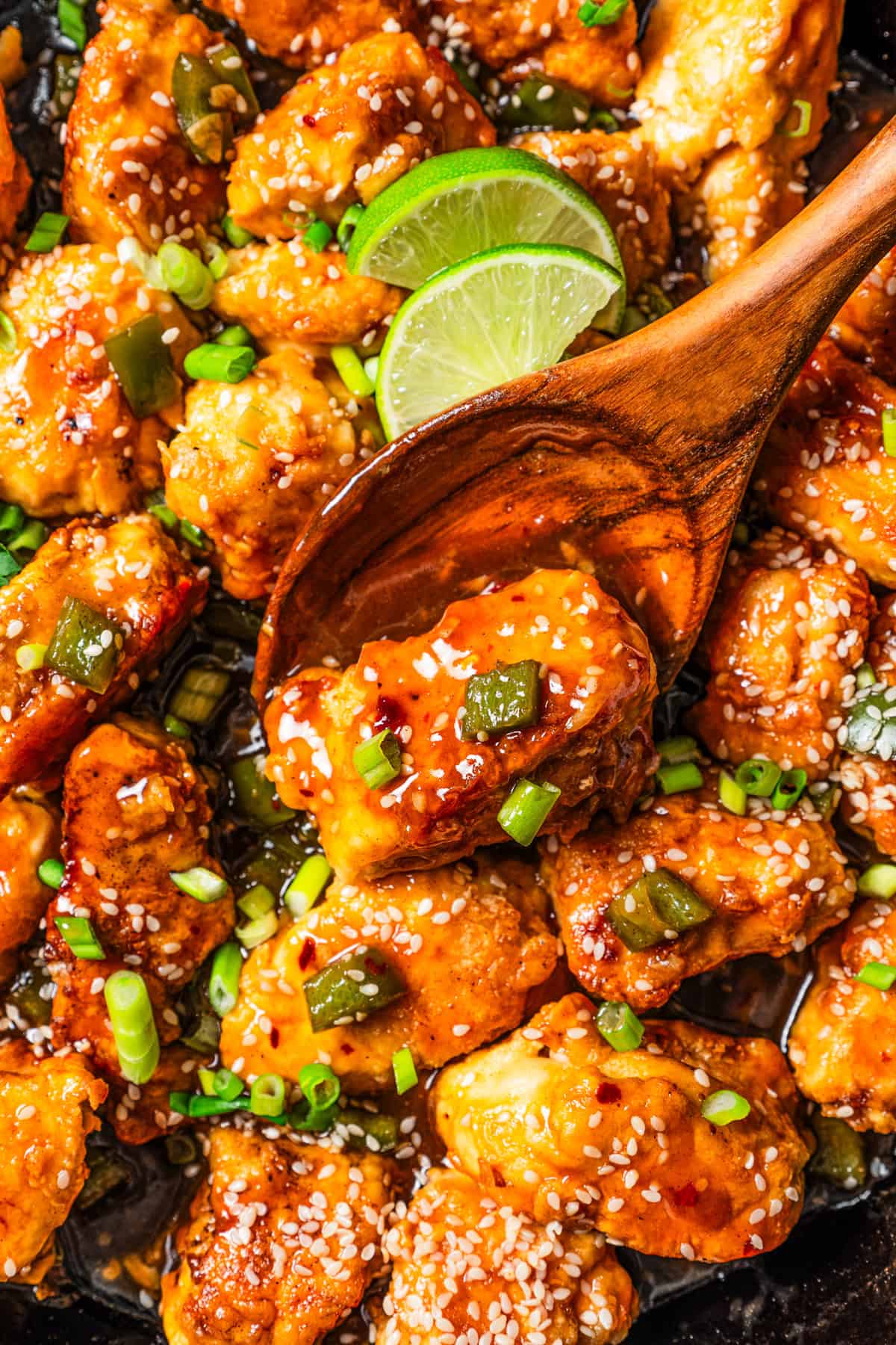 Close up of empress chicken in a skillet with a wooden spoon, garnished with lime wedges, sesame seeds, and green onions.