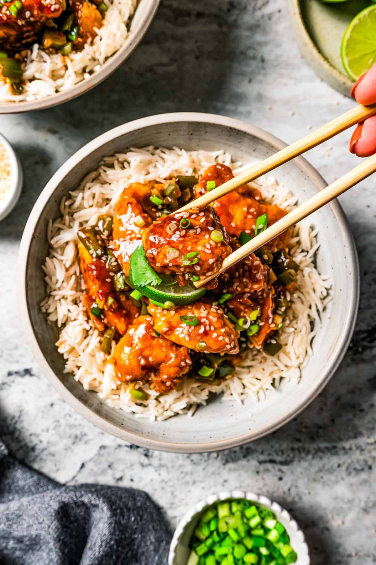 A pair of chopsticks holding a piece of chicken above a bowl of rice.