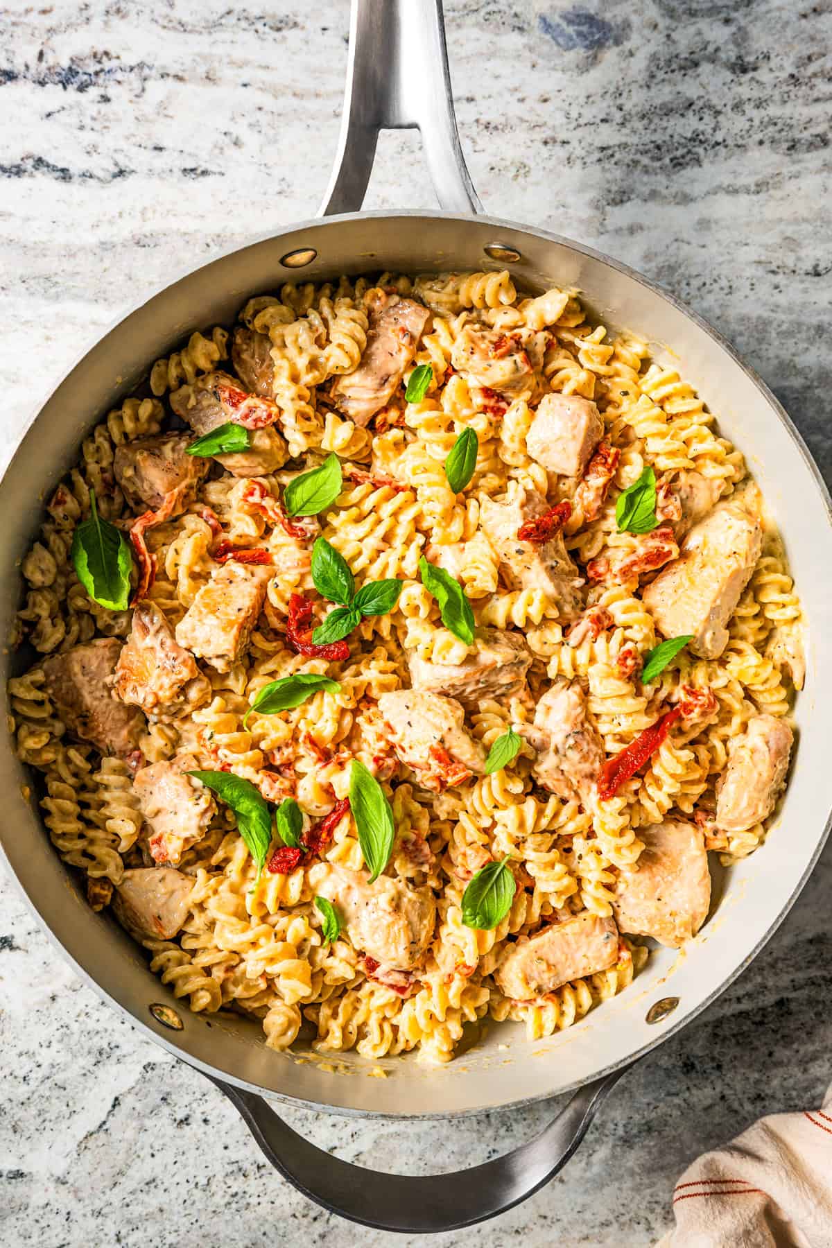 Bites of chicken and cooked pasta in a skillet garnished with basil leaves.