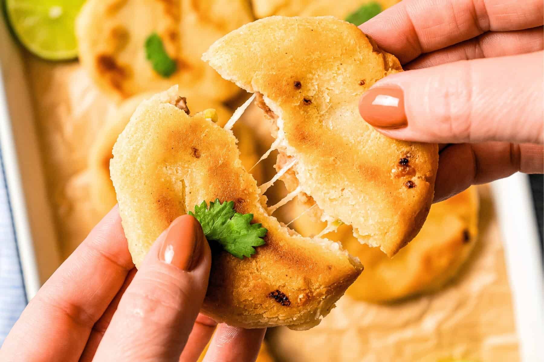 This Salvadoran pupusas recipe makes crispy masa cakes stuffed with refried beans, pickled jalapeños, and melted cheese. Served with curtido! Two hands breaking apart a pupusa over more pupusas on a tray.