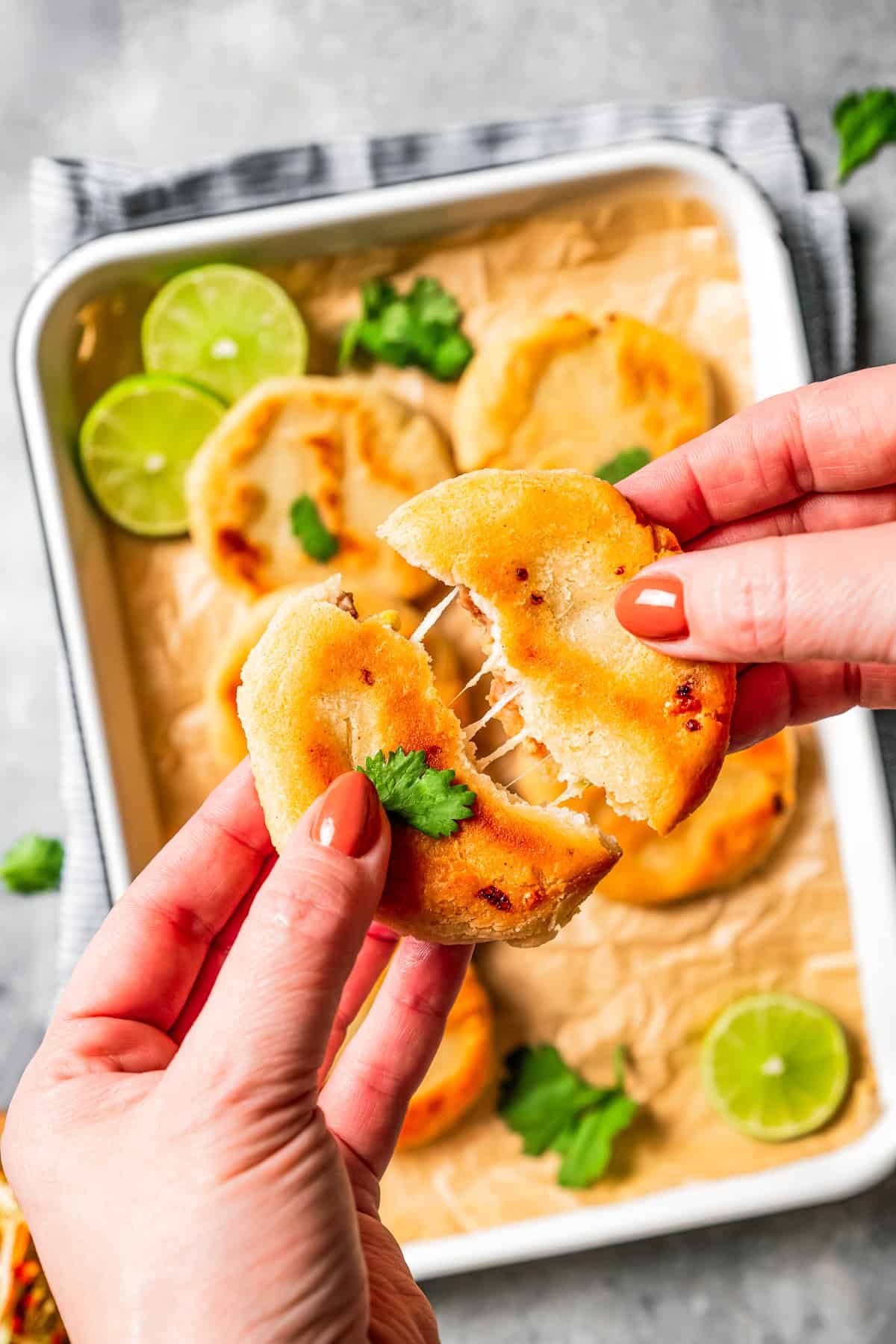 Two hands breaking apart a pupusa over more pupusas on a tray.