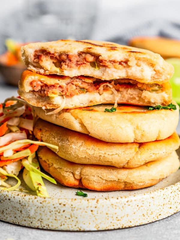 Pupusas stacked next to curtido on a stoneware plate with the top pupusa broken in half to reveal the refried bean filling inside.