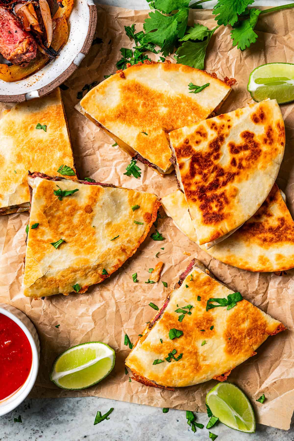 Overhead view of steak quesadillas on a sheet of parchment paper next to a bowl of salsa and lime wedges.