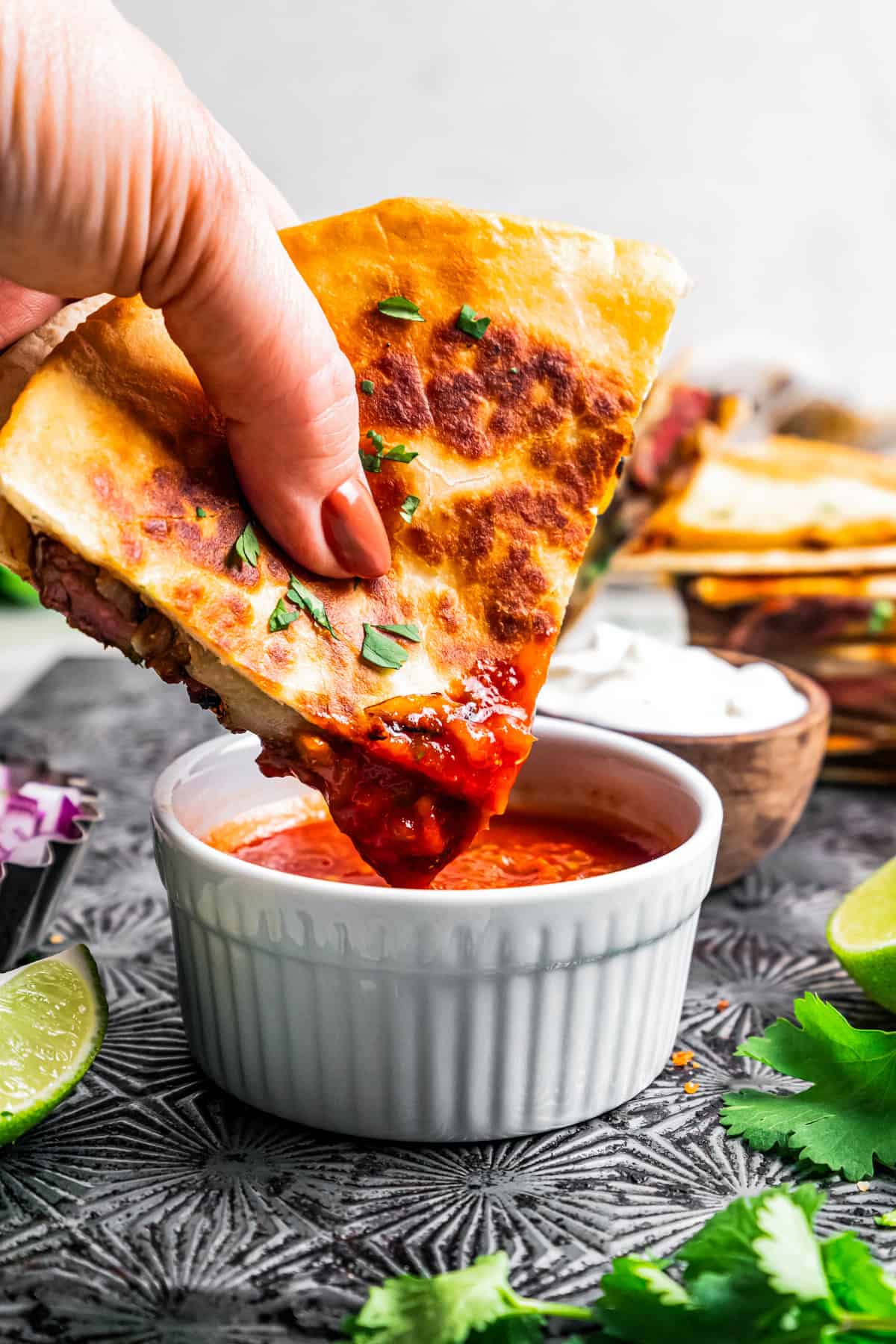 A hand dipping a steak quesadilla into a small bowl of salsa.