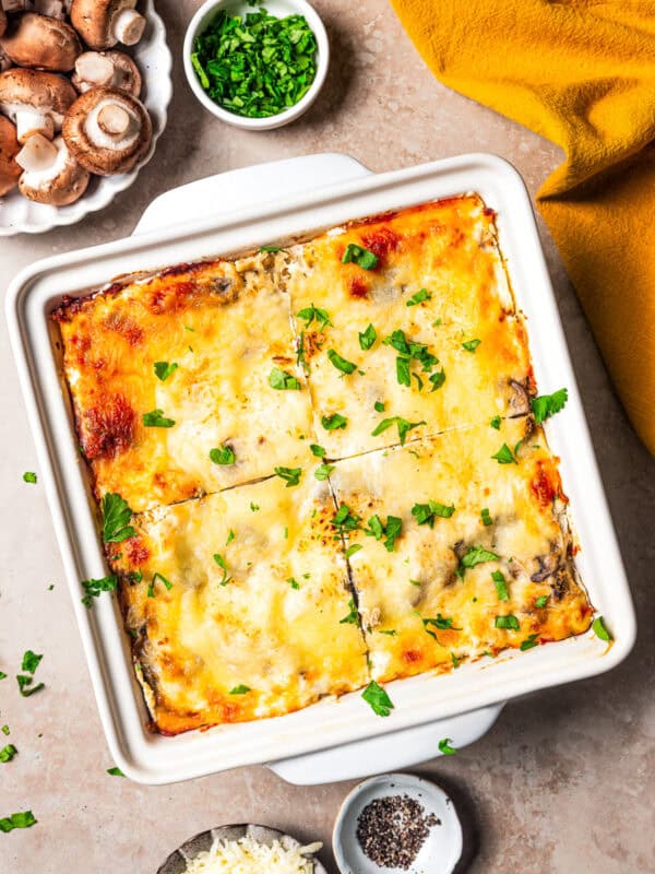 Whole lasagna cut into quarters in a ceramic baking dish, surrounded by bowls of ingredients.