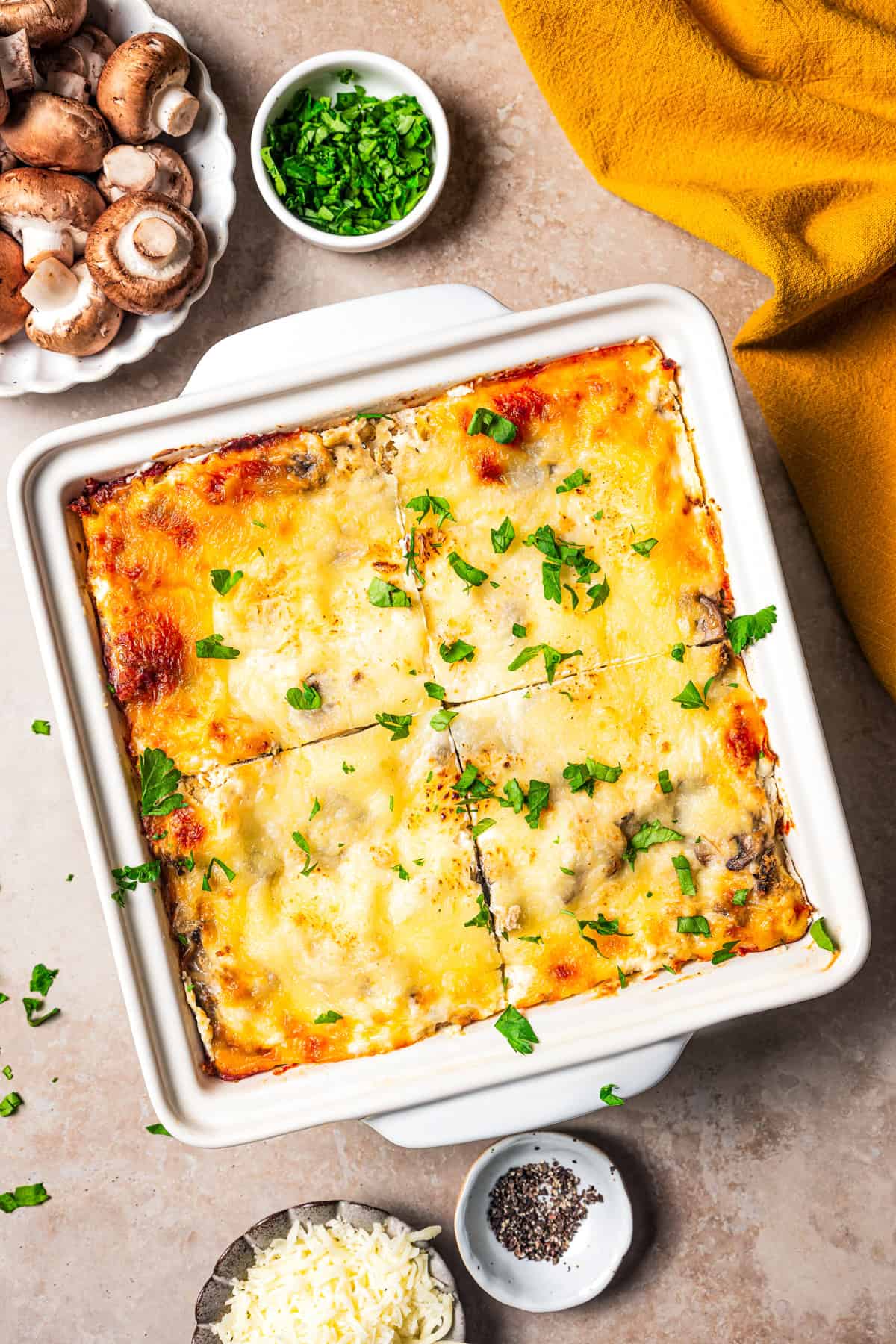 Whole lasagna cut into quarters in a ceramic baking dish, surrounded by bowls of ingredients.