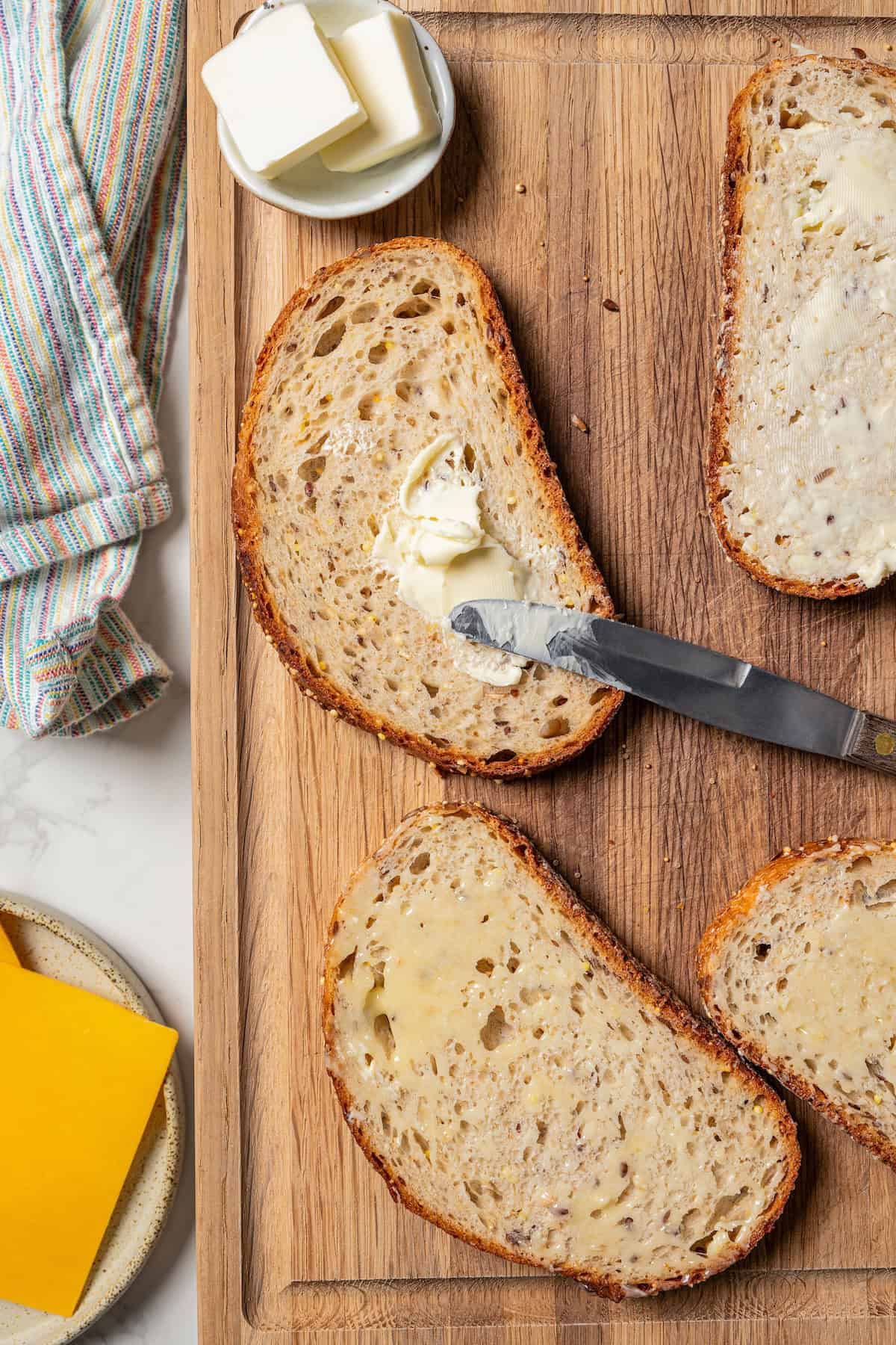 A knife spreading butter over a slice of bread, next to more bread slices on a wooden cutting board.