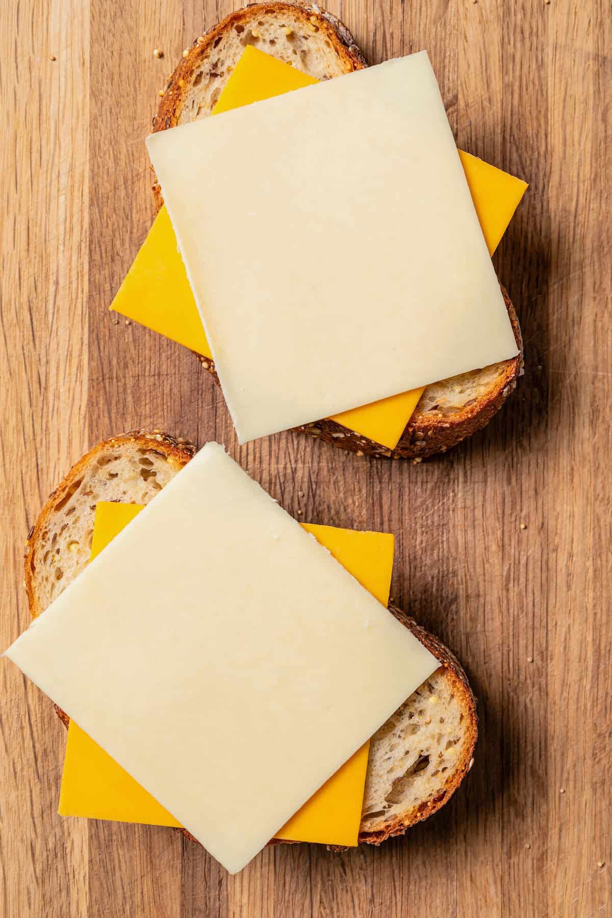 Slices of cheddar and Gruyere cheese layered over two bread slices on a wooden cutting board.