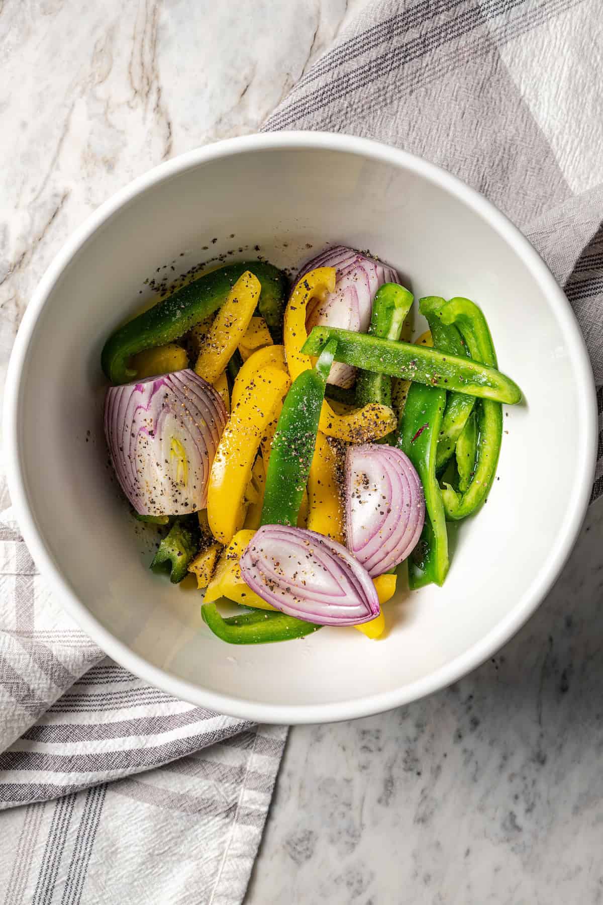 Raw sliced bell peppers and onions tossed with seasonings in a bowl.