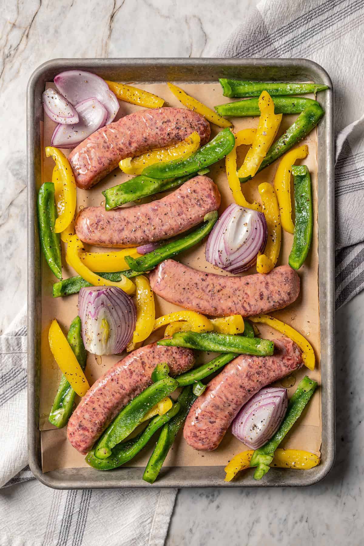 Seasoned bell peppers and onions spread on a baking sheet with Italian sausages.