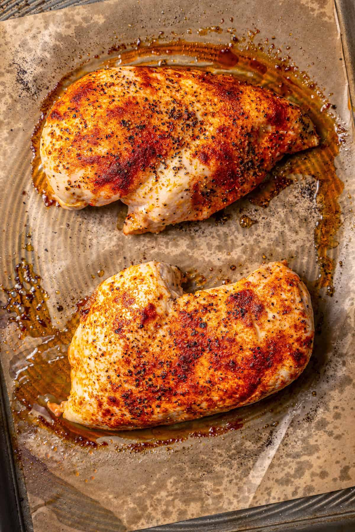 Two baked seasoned chicken breasts on a parchment-lined baking sheet.