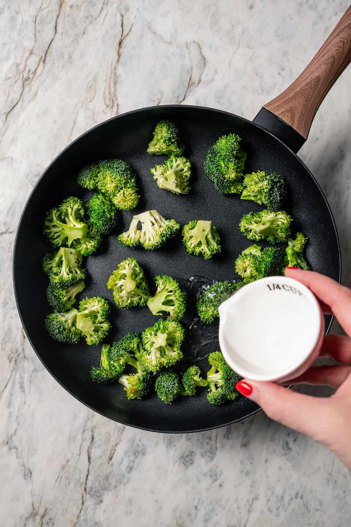 A hand pouring a small cup of water into a skillet with broccoli.