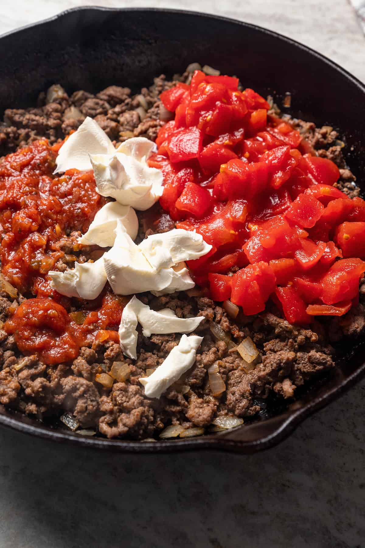 Tomatoes, salsa, and cream cheese added to browned ground taco meat in a skillet.