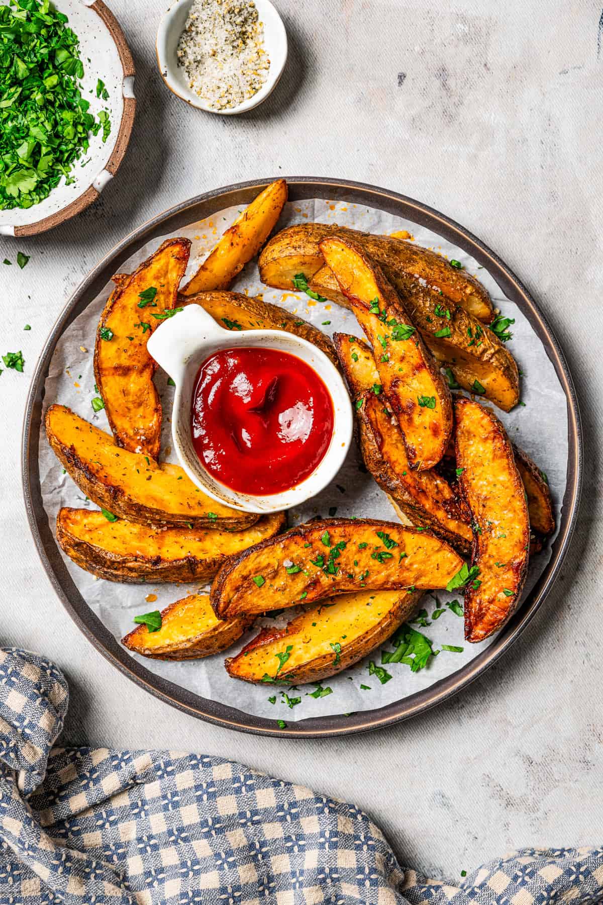 A plate with air fryer potato wedges, ketchup, and fresh parsley, with small bowls of parsley and seasoning in the background.