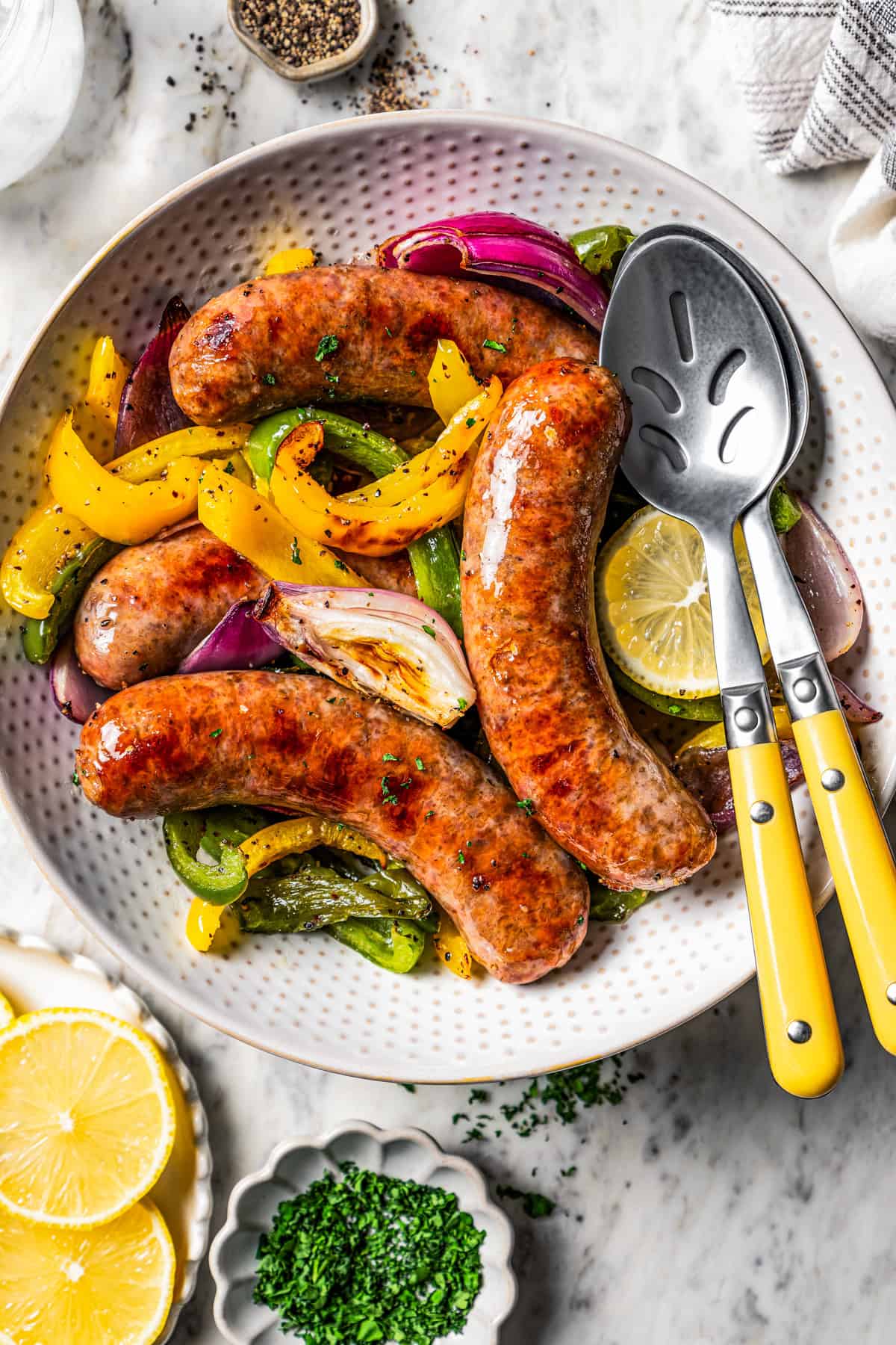 Oven-baked Italian sausage is an easy way to cook sausages that are juicy and caramelized, complete with a side of roasted peppers! Italian sausage and peppers served in a bowl with two serving spoons.